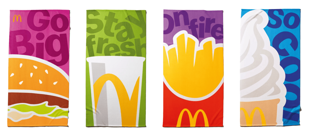 Due teli mare McDonald's con design colorati: uno raffigura un hamburger e l'altro una bibita, con frasi motivazionali sullo sfondo.